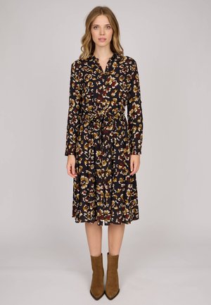 Une femme se tient de face, portant une robe midi à manches longues noire à fleurs, avec une taille nouée et des bottes ankle en daim marron.