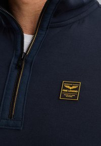Sudadera con cremallera azul marino hecha de un tejido texturizado, que presenta un parche con acentos amarillos que dice "PME Legend" y un cierre delantero con cremallera.