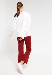 Weißer Kabelstrickpullover kombiniert mit roten Maßanzughosen und weißen Sneakern. Der Pullover hat ein strukturiertes, gemütliches Muster und gerippte Bündchen.