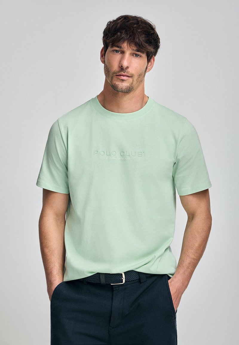 Mintgrön kortärmad t-shirt med präglad "POLO CLUB"-logotyp på framsidan. Tyget verkar mjukt; modellen bär mörka byxor och ett vävt bälte.