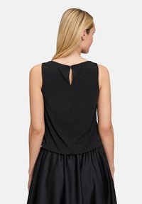 Schwarzes ärmelloses Oberteil mit leichter Textur und Keyhole-Rückendetail. Der Stoff wirkt leicht und glatt und ist mit einem voluminösen schwarzen Rock kombiniert.