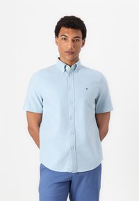 Chemise à manches courtes bleu clair en coton, avec un petit logo sur la poitrine, col standard et ourlet droit.