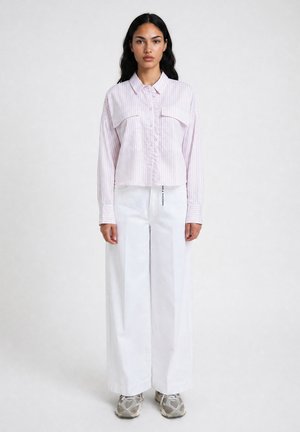 Woman with long dark hair wearing a light pink striped button-up shirt, wide white pants, and gray athletic shoes standing against a plain background.