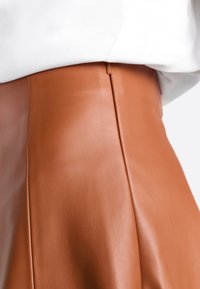 Gonna in finta pelle marrone con una texture liscia, dotata di un dettaglio cucito sul retro e un design aderente, abbinata a una maglietta bianca.