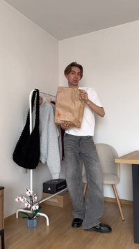 Bolsa de papel marrón con el texto "Ámame. Vísteme", jeans de pierna ancha grises, camiseta blanca, bolsa de punto negra y un perchero en una habitación neutra.