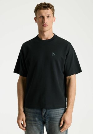 Young man with short curly hair wearing a black short-sleeve T-shirt and blue jeans, standing against a plain light background.