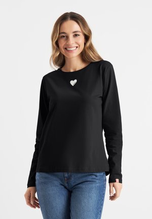 Zwarte longsleeve met ronde hals en een hartvormig accent in het wit. Draagt een blauwe denimjeans. Eenvoudige, gladde textuur.