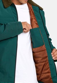 Giacca esterna verde acqua con colletto in pelliccia, aperta per rivelare la fodera interna color rame e una tasca. Sotto, una maglietta bianca semplice.