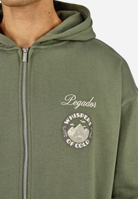 Olajzöld cipzáros kapucnis pulóver, puha anyagból készült, elöl logo folttal, amely hegyeket és a "Cold Suttogása" szöveget ábrázolja.