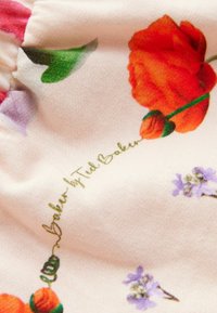 Kwiatowa tkanina na kremowym tle, z czerwonymi różami i fioletowymi kwiatami, mająca teksturowaną powierzchnię i złotym napisem "Ted Baker".