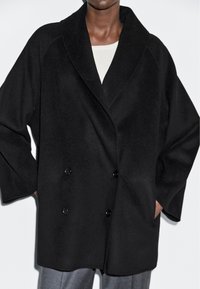 Zwarte dubbelknopenjas met een brede revers, gemaakt van een gestructureerde stof. Beschikt over zijzakken en een relaxte pasvorm.