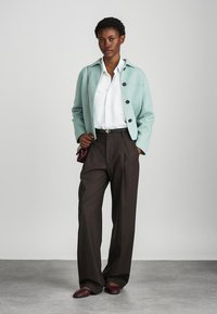 Světlá modro-zelená bunda s knoflíky, bílá košile, hnědé kalhoty a vínové boty. Oblečení se vyznačuje strukturovaným designem a na míru střihem.