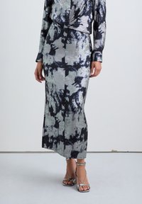 Nainen, jolla on pitkä musta-harmaa tie-dye-kuvioinen mekko, jossa on pitkät hihat ja hopeiset korkeat sandaalit.