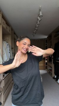 T-shirt nero indossato da una persona con pelle chiara, sorridente, con le braccia sollevate. Lo sfondo presenta mobili in legno chiaro per la conservazione e vestiti.