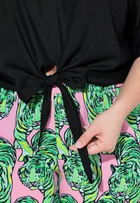 Top corto nero annodato abbinato a pantaloni corti rosa con stampe di tigri verdi e vita elasticizzata. Primo piano sul nodo e sul dettaglio della mano.