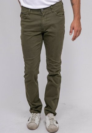 Homme portant un pantalon slim vert olive et des baskets blanches, debout avec une main détendue le long du corps, sur un fond uni.