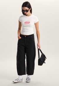 Camiseta blanca ajustada con gráfico rojo, pantalones negros de pierna ancha, zapatos plateados y bolso negro. Los accesorios incluyen gafas de sol oscuras.