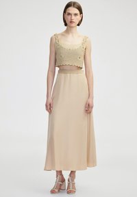 Beige gehaakte crop top met golvende randen en bloemdetails, gecombineerd met een vloeiende, enkel-lange beige rok en lichte sandalen met bandjes.