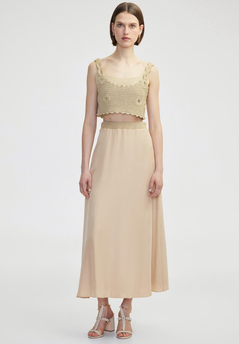 Top corto de ganchillo beige con bordes en forma de ondas y detalles florales, combinado con una falda larga beige y fluida hasta los tobillos y unos tacones ligeros con tiras.