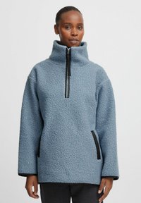 Világoskék fleece pulóver magas nyakkal, félcipzárral elöl, oldalzsebekkel és fekete részletekkel a cipzárnál és az alján. Texturált szövetfelület.