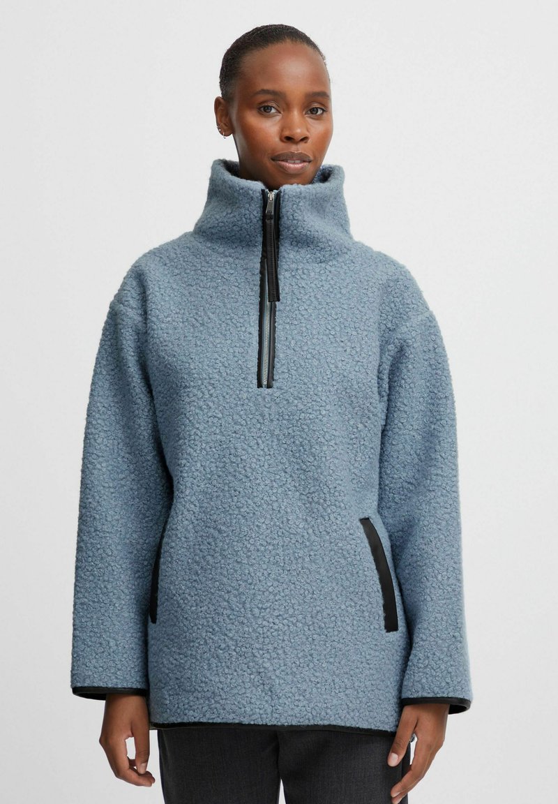 Világoskék fleece pulóver magas nyakkal, félcipzárral elöl, oldalzsebekkel és fekete részletekkel a cipzárnál és az alján. Texturált szövetfelület.