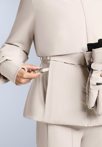 Bež vanjska odjeća s uskim krojem, s sigurnosnim pojasom i bočnim džepom. U drugoj ruci drži se par sivih rukavica s crnim detaljima.