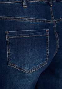 Jeans de mezclilla azul oscuro con un bolsillo trasero con costura naranja, textura vertical y un ligero desvanecimiento cerca de los bordes.