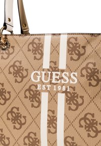 Beige handbag with a textured pattern featuring the "GUESS" logo in white, accented by two vertical white stripes and a circular hardware detail.