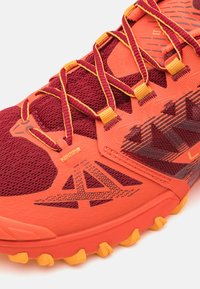 Rode en oranje sportieve schoen met een gestructureerde gebreide bovenkant, contrasterende veters, rubberen accenten en een patroon aan de zijkant; ontworpen voor grip en stabiliteit.