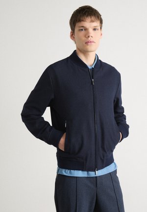 Marinefarbene Bomberjacke aus weichem Material mit Reißverschluss, gerippten Bündchen und Seitentaschen, kombiniert mit einem hellblauen Hemd darunter.