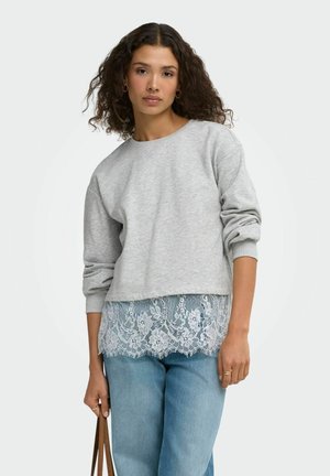 Femme aux cheveux bouclés portant un sweat-shirt gris superposé à un haut en dentelle blanche, un jean bleu, et tenant une sangle de sac marron dans une main.