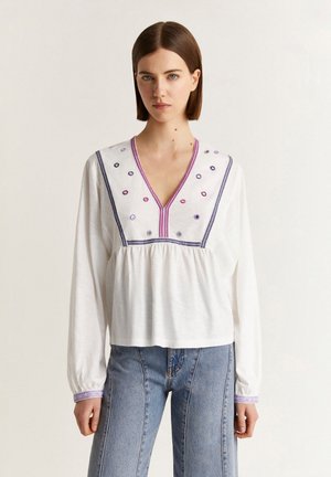 Camicia bianca a maniche lunghe con scollo a V, caratterizzata da un ricamo circolare colorato e una vita arricciata. Dettagliata con cuciture viola sui polsi.