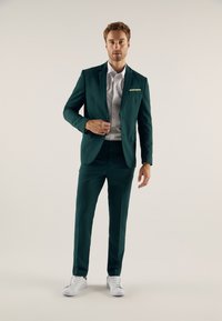 Tiqui Suit mit schmaler Passform, ausgestattet mit einer Brusttasche mit gestreiftem Akzent, kombiniert mit einem weißen Hemd und weißen Sneakern.