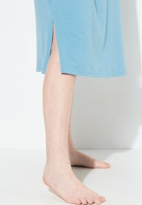 Ljusblå bomullsklänning med sidoklyftor, kort längd och mjuk yta, visas från sidan med barfota synligt.