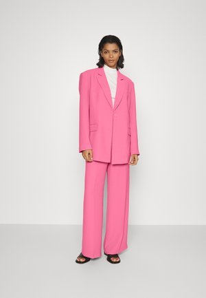 Blazer oversized fúcsia com calças de perna larga a condizer. Textura suave, fecho com um botão único e dois bolsos frontais. Modelo a usar sand�álias pretas.