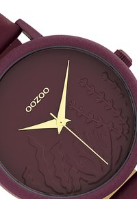 Montre ronde de couleur marron avec un cadran texturé mettant en avant des motifs de plantes aquatiques, des aiguilles des heures et des minutes dorées, ainsi qu'un logo doré simple. Bracelet en silicone.