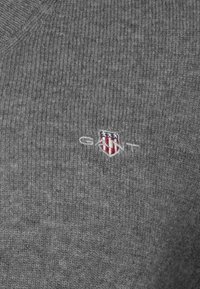 Maglione grigio lavorato a maglia con texture a coste, caratterizzato da un piccolo logo ricamato in rosso, bianco e blu sul lato sinistro.