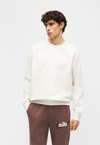 Νεαρός άνδρας με σκούρα σγουρά μαλλιά, φορώντας λευκό φούτερ και καφέ παντελόνι Nike, με τα χέρια στις τσέπες, κοιτάζει αριστερά.