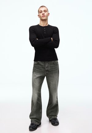 Giovane uomo in piedi con le braccia incrociate, indossa una maglia nera a maniche lunghe, jeans grigi larghi e scarpe da ginnastica nere, su uno sfondo bianco uniforme.