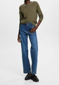 Femme portant un pull en tricot vert olive, un jean bleu taille haute, des mocassins noirs et des bagues en argent, se tenant avec une main dans la poche.