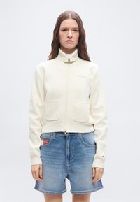 Γυναίκα με μακριά καστανά μαλλιά που φοράει εκρού ζακέτα φλις με φερμουάρ και ανοιχτόχρωμα τζιν σορτς, στέκεται μπροστά σε απλό φόντο.