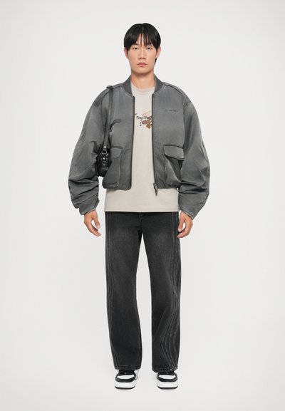 Gray bomber jacket with voluminous sleeves, two front pockets, worn over a cream T-shirt; paired with dark jeans and black sneakers.