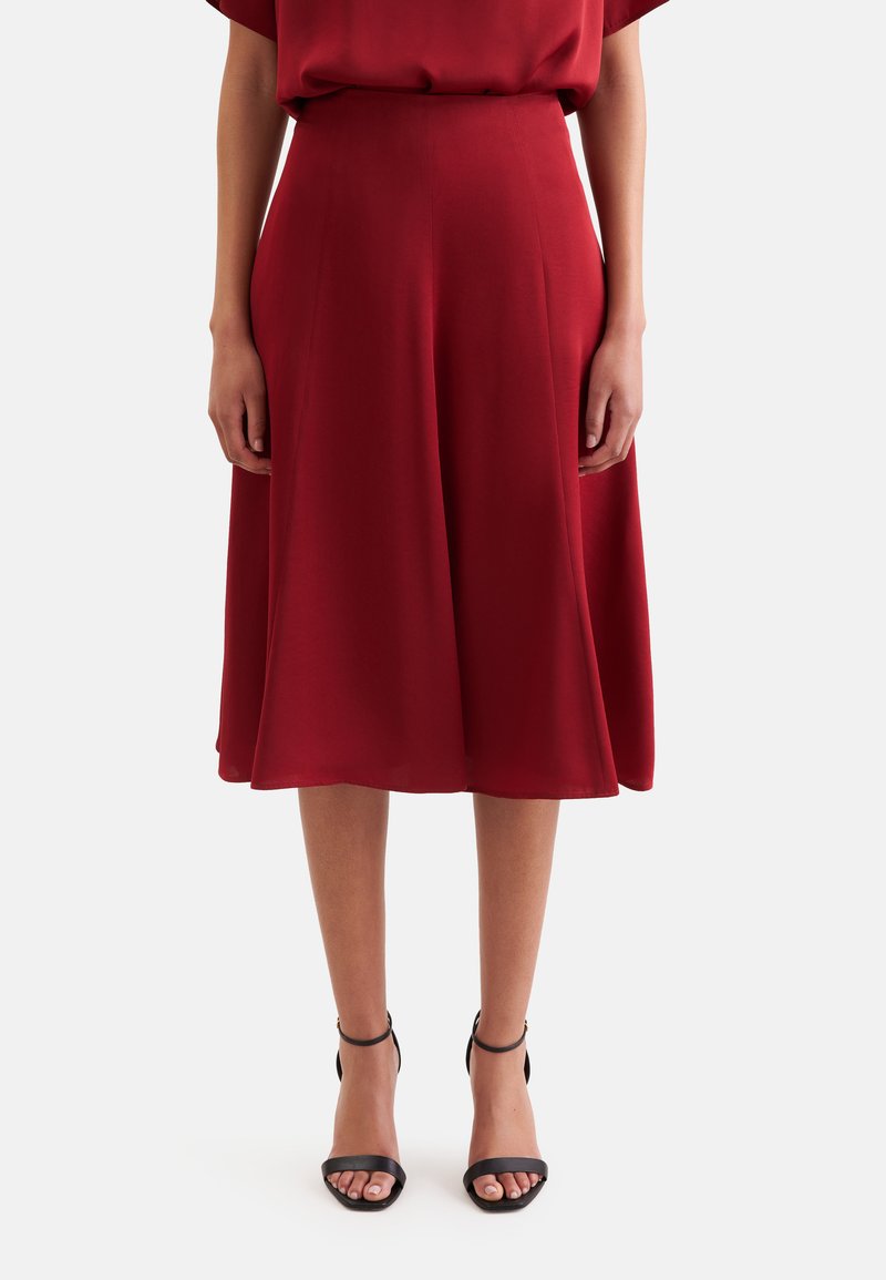 Femme portant une jupe rouge taille haute jusqu’au genou, associée à des talons noirs ouverts avec bride à la cheville, debout devant un fond uni.