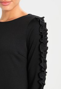 Femme portant un haut noir à manches longues avec des détails froncés le long de l'épaule et du bas de la manche, sur fond uni.