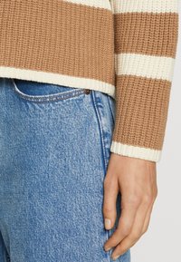 Mano descansando a un lado, vistiendo jeans de mezclilla azul y un suéter de punto a rayas beige y blanco.