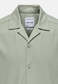 Camisa de canalé verde claro con cuello, que presenta dos botones y una etiqueta interior. Textura suave, diseño de corte relajado.
