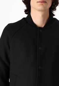 Svart ullblandad bomberjacka med ribbat krage, tryckknappsstängning och sidofickor; har en slät, strukturerad yta.