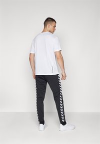 Witte T-shirt met korte mouwen, gecombineerd met zwarte sportbroek met witte chevronstrepen langs de zijkanten en witte sneakers.
