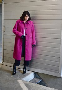 Fuksiaroosa ülikondne mantel tekstuuriga kangast, millel on krae, mustad nööbid ja küljetaskud, kantud mustade pükste ja ankle'ides jalatsitega.