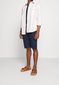 Camicetta a maniche corte a righe in colori chiari, pantaloni cargo blu navy e sandali marroni con fibbia, che mostrano un look estivo informale.